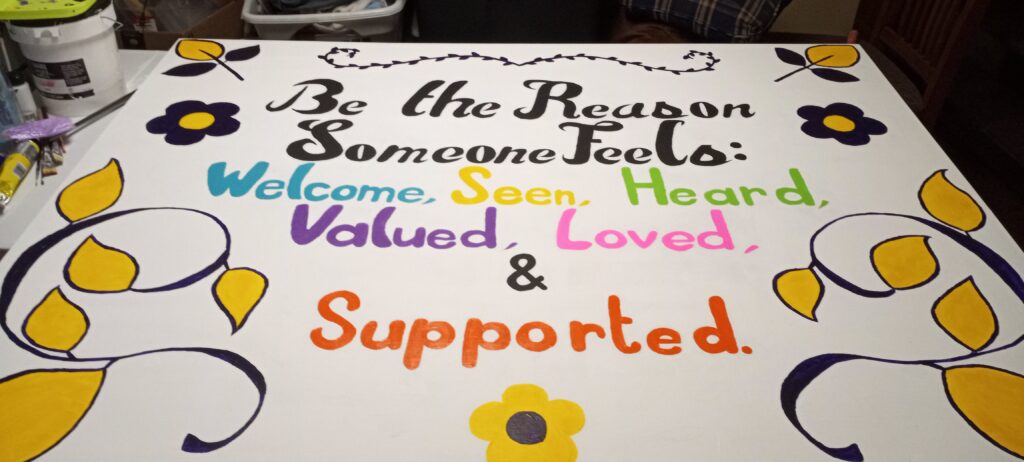 Typography painting.There is some slight floral filigree in purple and gold that surround the words: "Be the Reason Someone Feels: Welcomed, Seen, Heard, Valued, Loved, & Supported." The last 6 words are in blue, yellow, green, purple, pink, and orange respectfully.